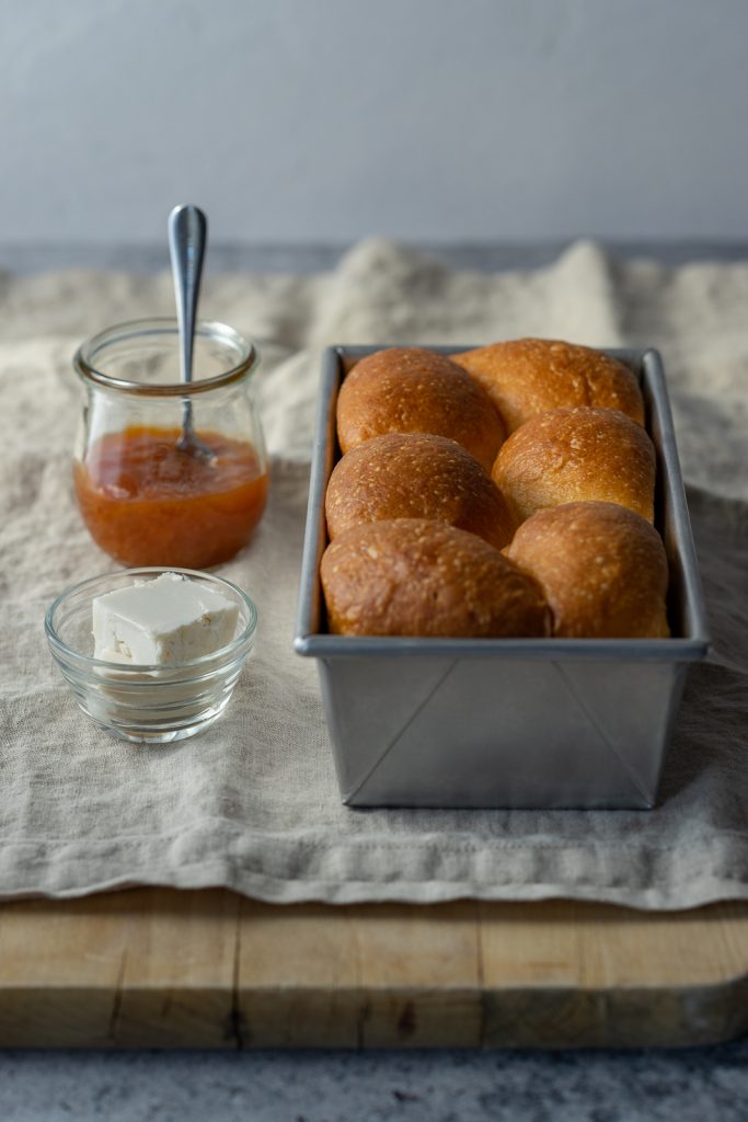 A golden brown brioche is in a bread loaf pan and is fresh out of the oven. To the left of the pan is a small jar of apricot jam with a spoon in the jar, inviting you to smother the bread in jam. There are two small pats of bread in a small bowl directly in front of the jam. All of the objects are resting on a linen napkin on top of a wooden cutting board.