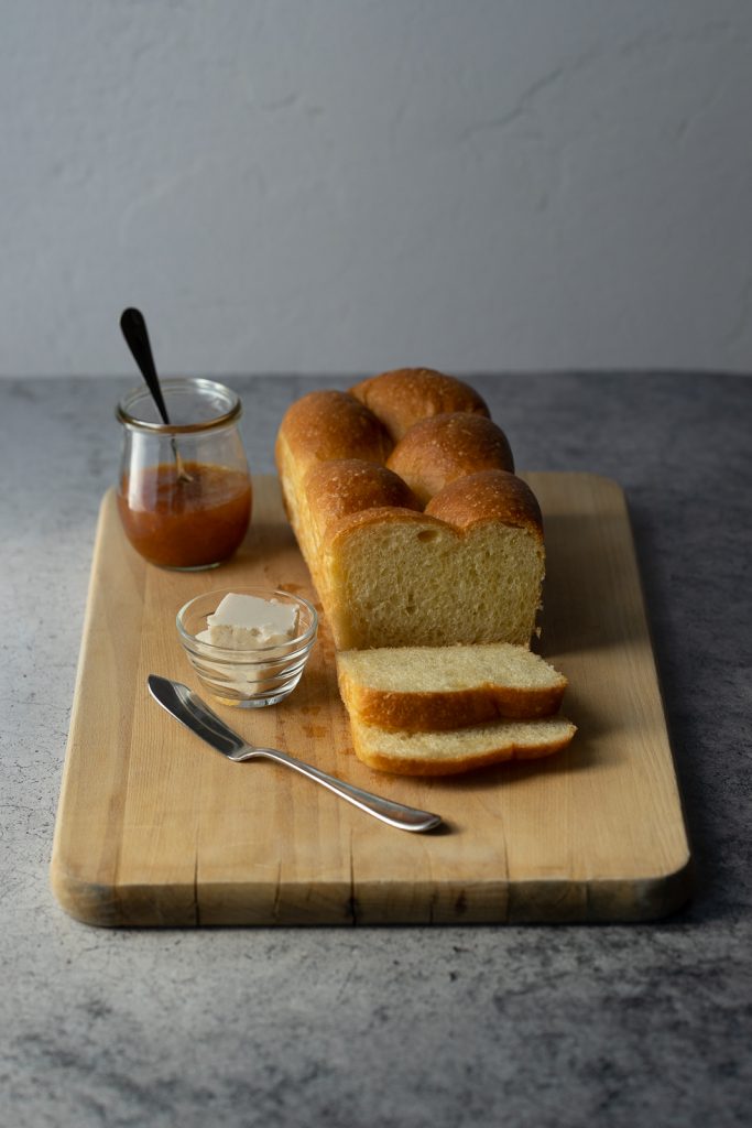 An image of a vegan brioche loaf two pieces sliced off of the end sitting in front of the loaf. A butter serving utensil is to the left of the bread in the foreground.