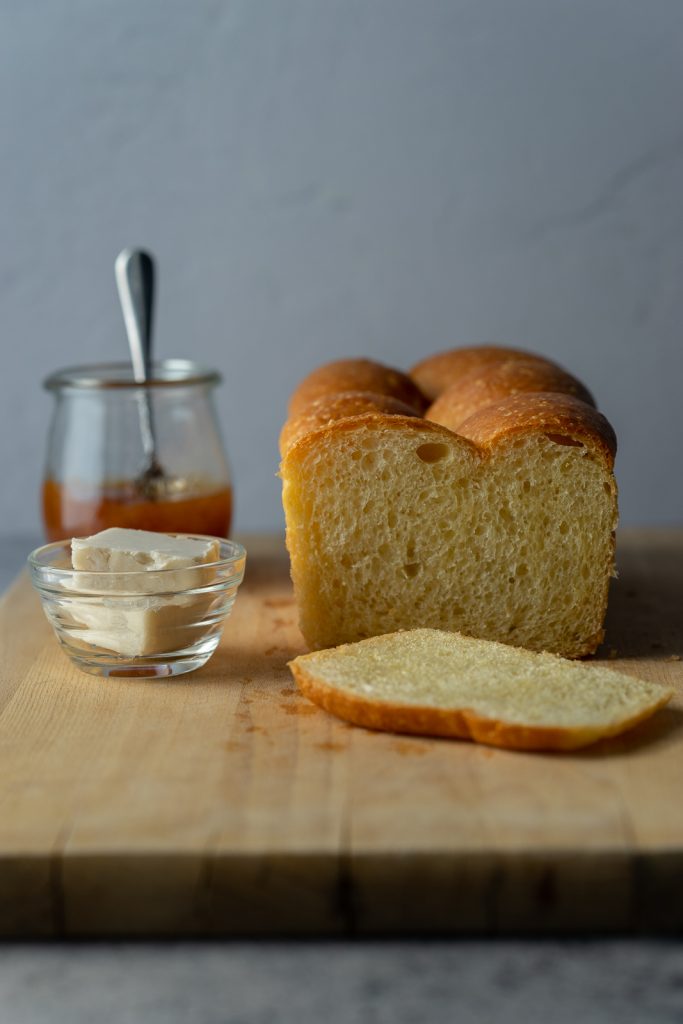 A close up image of a vegan brioche loaf with the end sliced off. A small dish of butter and apricot jam sit to the left of the loaf.