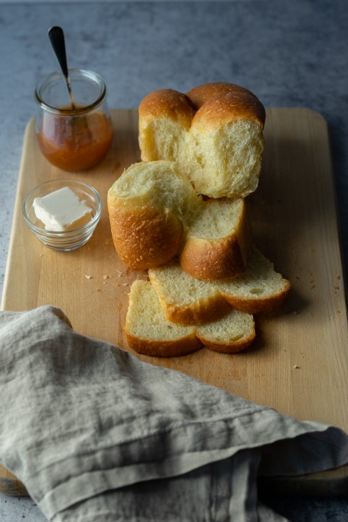 An image of a vegan brioche loaf torn apart to reveal the insides. A small dish of butter and apricot jam sit to the left of the loaf. A linen napkin is in the foreground.