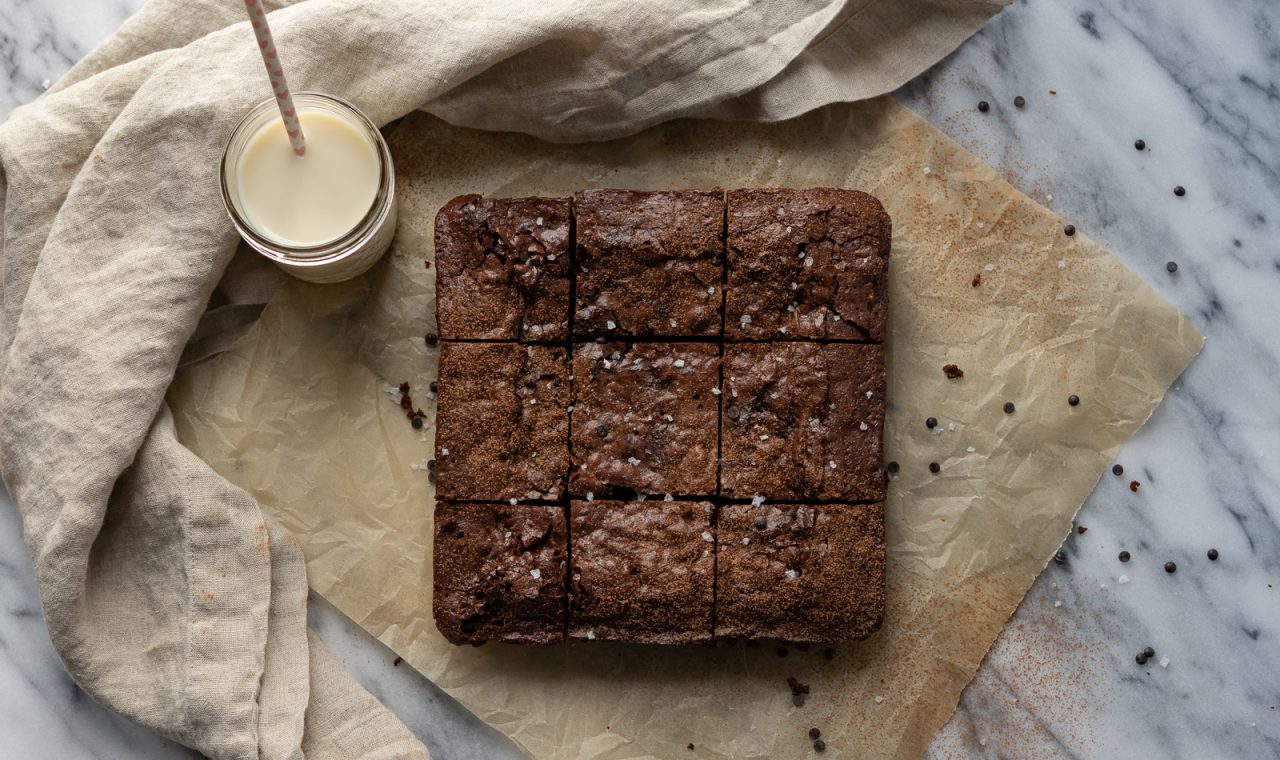 Nine squares of brownies are sitting atop a square of parchment paper. A glass of milk with a straw sits to the upper left corner and a cloth napkin frames the whole image.