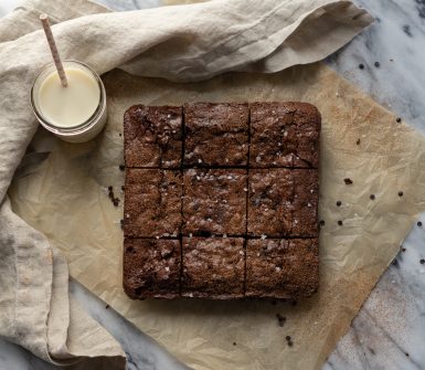 Nine squares of brownies are sitting atop a square of parchment paper. A glass of milk with a straw sits to the upper left corner and a cloth napkin frames the whole image.