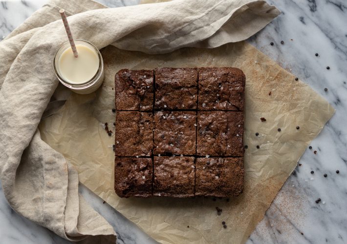 Nine squares of brownies are sitting atop a square of parchment paper. A glass of milk with a straw sits to the upper left corner and a cloth napkin frames the whole image.