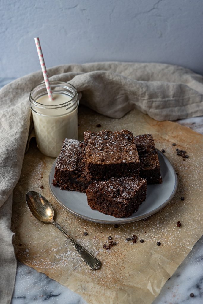 A small plate of four brownies squares are sitting atop a square of parchment paper. A glass of milk with a straw sits to the upper left corner and a cloth napkin frames the whole image. There is an antique spoon in the lower left corner.