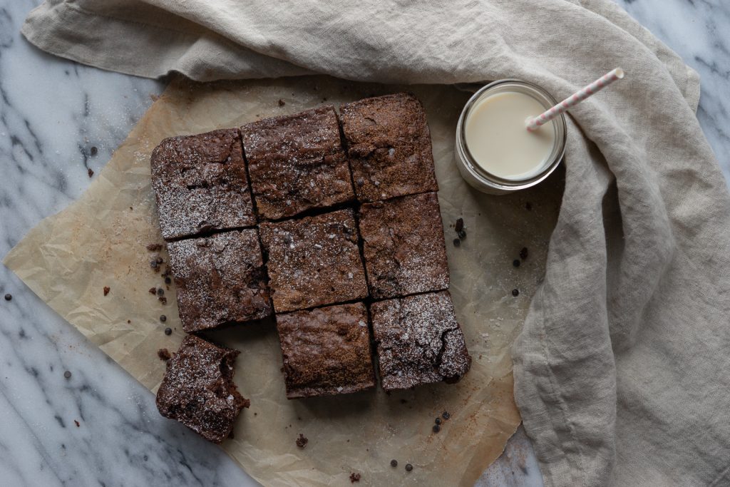 Nine squares of brownies are sitting atop a square of parchment paper. A glass of milk with a straw sits to the upper right corner and a cloth napkin frames the whole image.