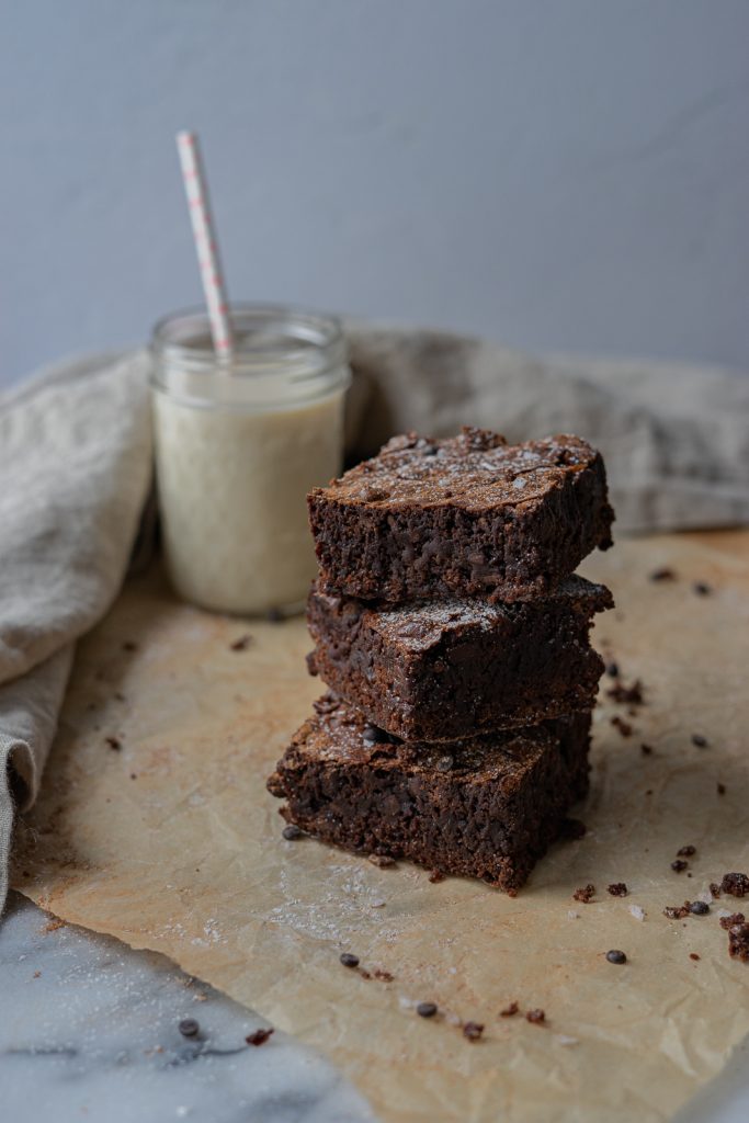 Three squares of brownies are stacked atop of each other. A glass of milk with a straw sits to the upper left corner and a cloth napkin frames the whole image.