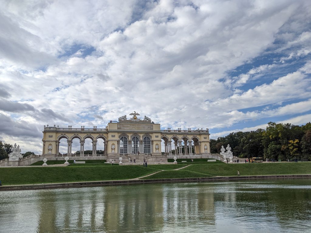 The Gloriette building on the grounds of the Schönbrunn Palace in Vienna, Austria. In front of the building is a pond and there are many white, fluffy clouds in the sky above the building. There are green trees to the right of the building.  