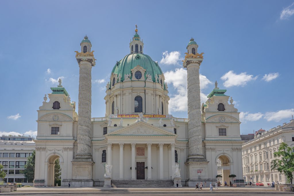 St. Charles Church, a Catholic Church in Vienna, Austria. There is one main dome that is aquamarine green in color due to the patina. There are two large columns on either side of the main dome with mini domes that are also patinaed. At the top of the columns, underneath the domes are gold-leafed animals. 