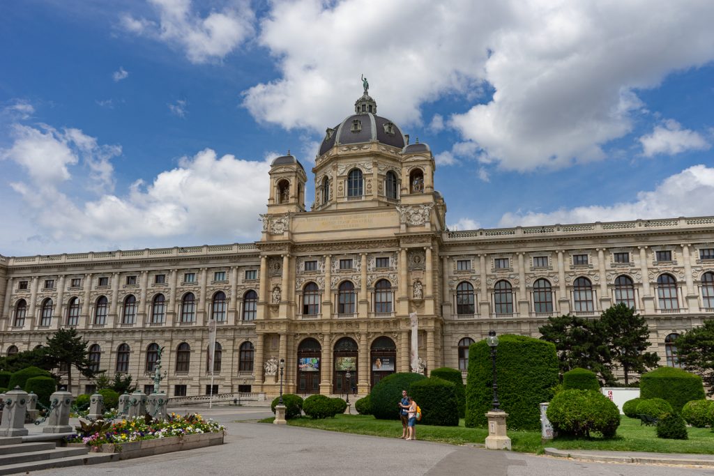 The natural history museum in Vienna, Austria. The building has a main entryway that is taller than the rest of the building. There is a central dome that is dark grey in color with four smaller domes on each corner surrounding the main dome. There are two tourists standing in front of the building.