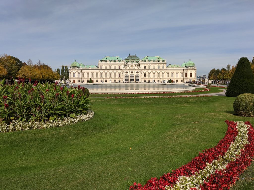 The Schloss Belvedere upper building. The whole roof of the building is patinaed and there are four domes on each corner of the building.  In front of the palace is a large man-made pond and the grounds surrounding the pond are well kept. There are red and white flowers in the lower right front corner of the picture.