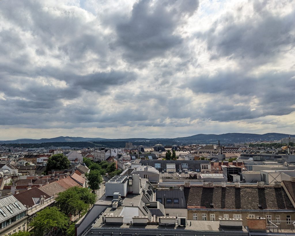 A view from a rooftop deck of the city of Vienna, Austria just before a rain storm is about to hit. The clouds are darkened on the bottom, but light at the top, as the sun is still shinning behind them. 