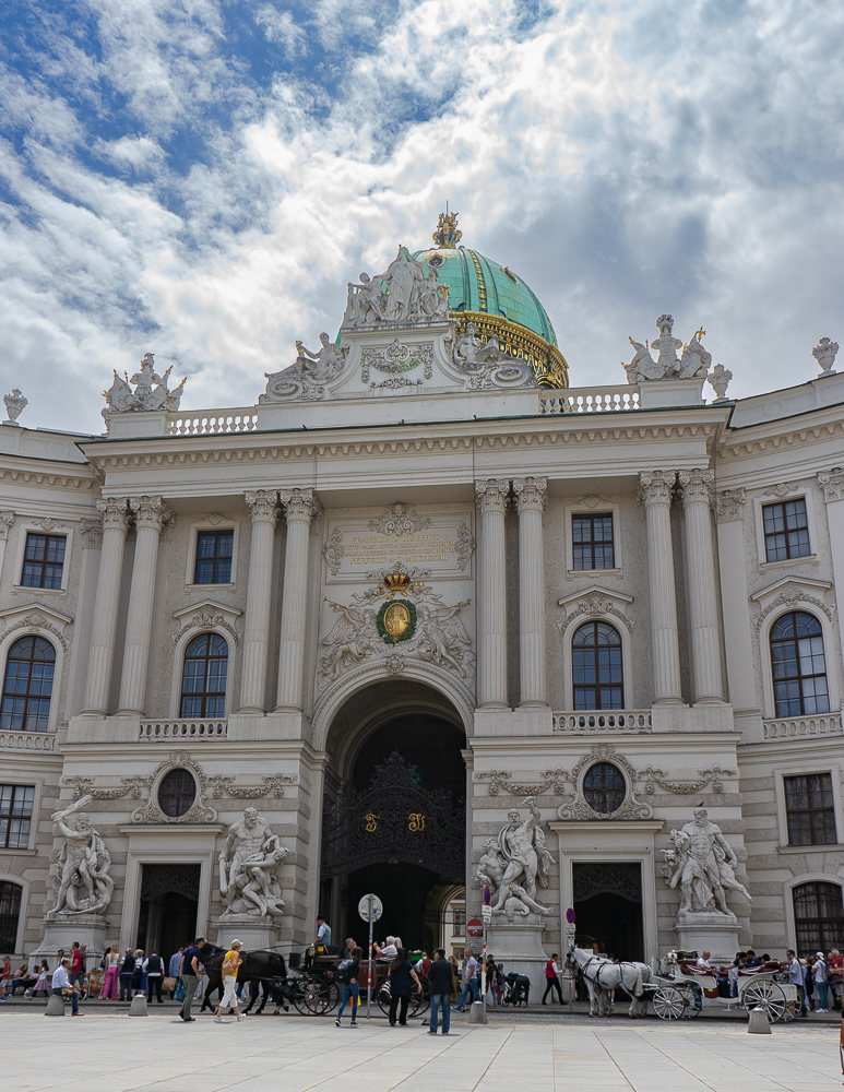 Hofburg Palace in Vienna in the summer. There are fluffy clouds in the sky above the building. The dome is aquamarine green due to patina with gold trim. There are four statues at the opening of the palace and four sets of two columns above the head's of the statues. There are two horse drawn carriages in front of the building.