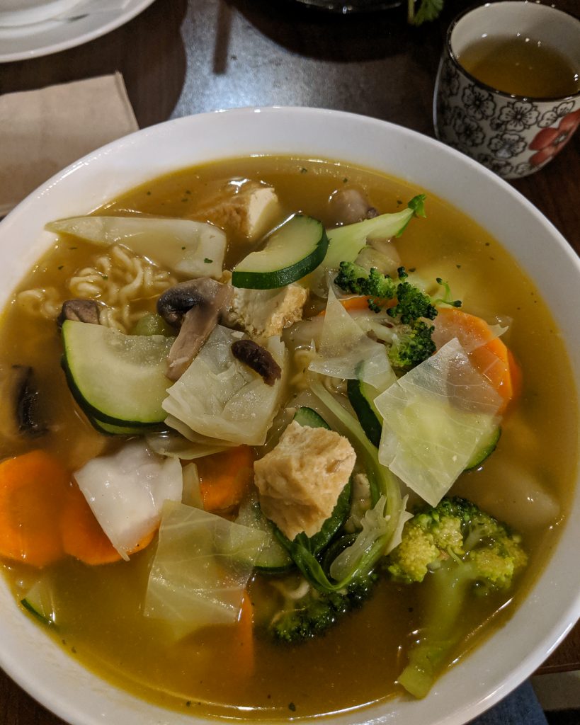 A bowl of soup from a Vietnamese restaurant. The soup is a broth based soup with a mixture of vegetables and tofu. There is a cup of green tea in the upper right corner of the photo.