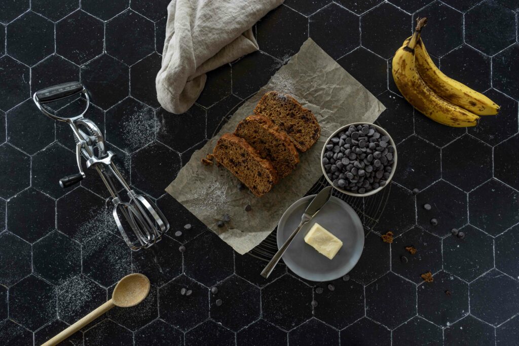 At the center of the image, there are three slices of vegan chocolate chip banana bread. There are two bananas, a pat of butter, and a small bowl of chocolate chips to the right of the bread. To the left there is an old-fashioned hand mixer and a wooden spoon. The light is coming from the the top of the image, bathing everything and creating shadows on the foreground. There are chocolate chips and powdered sugar scattered randomly throughout the image. There is a cream colored napkin at the top of the image, framing the bread.