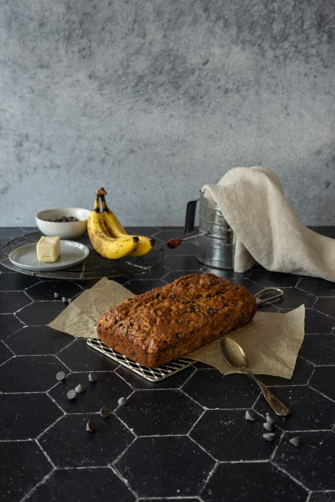 At the center of the image, there is a loaf of vegan chocolate chip banana bread that sits on top of a cooling rack. There is an old-fashioned hand flour sifter with a cream colored napkin inside of it, spilling over the edge, two bananas, a pat of butter, and small bowl of chocolate chips in the background. In the foreground, there is an antique spoon. The light is coming from the left side of the image, softly bathing everything and creating shadows on the right side of everything. There are chocolate chips scattered randomly throughout the image.