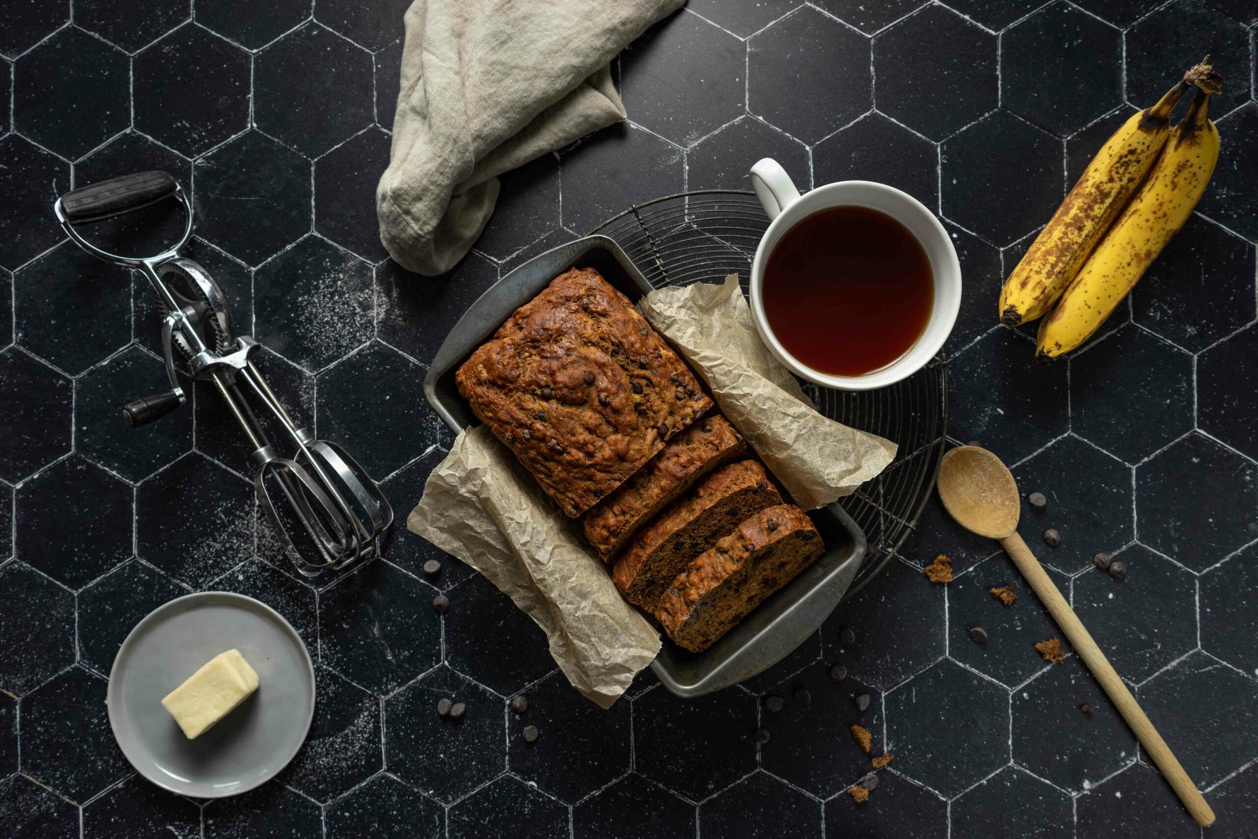 At the center of the image, there is a loaf of vegan chocolate chip banana bread that sits inside of the pan it was baked in. A cup of tea, a wooden spoon, and two bananas are to the right of the banana bread, and a small dish with butter and a hand mixer are to the left. Finally, there is a cream colored napkin to the top of the image. The light comes in from the top of the image by the napkin, and softly bends around the left side of the image, encircling the pan. There are chocolate chips scattered randomly throughout the image.