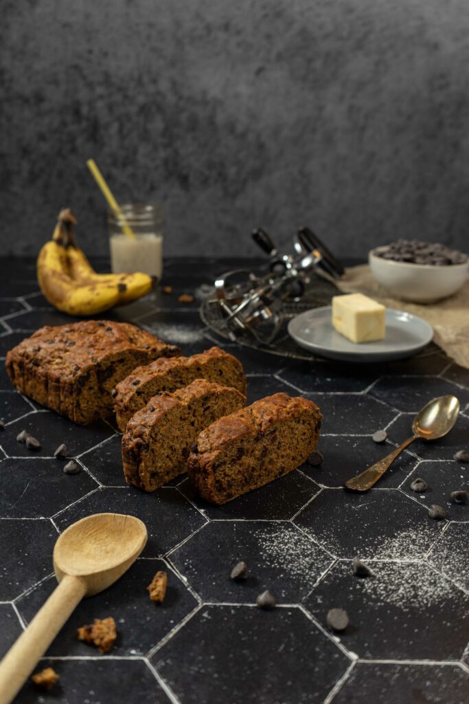 At the center of the image, there is a loaf of sliced vegan chocolate chip banana bread. There is an old-fashioned hand mixer, two bananas, a pat of butter, a small glass of milk with a straw, and a small bowl of chocolate chips in the background. In the foreground, there is an antique spoon and a wooden spoon. The light is coming from the left & right sides of the image, softly bathing everything and creating shadows on the foreground. There are chocolate chips and powdered sugar scattered randomly throughout the image.