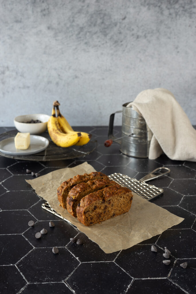 At the center of the image, there are three slices of vegan chocolate chip banana bread that sits on top of a cooling rack. There is an old-fashioned hand flour sifter with a cream colored napkin inside of it, spilling over the edge, two bananas, a pat of butter, and small bowl of chocolate chips in the background. The light is coming from the left side of the image, softly bathing everything and creating shadows on the right side of everything. There are chocolate chips scattered randomly throughout the image.